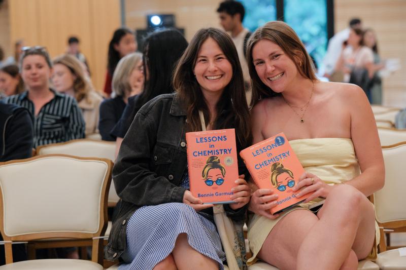 Two female audience members smiling and holding book titled "Lessons in Chemistry" by Bonnie Garmus